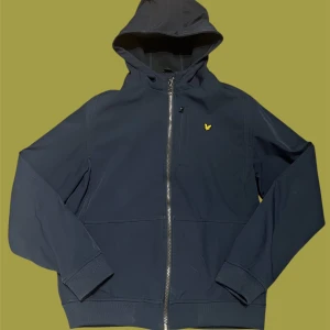 Mörkblå vindjacka från Lyle & Scott - Snygg mörkblå höst/vårjacka från Lyle & Scott med huva och dragkedja framtill. Jackan har ett gult logomärke på bröstet, ribbade muddar och två fickor. Perfekt för dig som vill ha en stilren och sportig look.