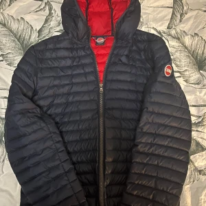 Mörkblå Colmar dunjacka med huva - Säljer en mörkblå dunjacka från Colmar med röd insida och huva. Jackan har quiltad design, dragkedja framtill och Colmar-logga på ärmen. Fylld med naturligt dun och har en clean, sportig look som funkar till streetwear. Perfekt för kalla dagar. Storlek 172CM 