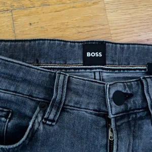 Grå Hugo Boss jeans slim fit - Snygga grå jeans från Boss med klassisk femficksdesign och svart Boss-lapp bak i midjan. Jeansen har rak passform och normal midja, tillverkade i mjukt jeanstyg med dragkedja och knapp framtill. Perfekta för dig som gillar stilrena och tidlösa jeans.