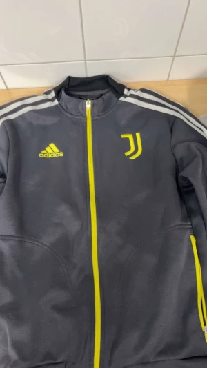 Grå Adidas Juventus track jacket - Grå track jacket från Adidas med Juventus-logga och gula detaljer. Jackan har hel dragkedja framtill, två fickor och klassiska vita ränder på axlarna. Perfekt för dig som gillar sportig stil och fotboll.