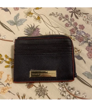 Svart plånbok från Tommy Hilfiger - Snygg svart plånbok från Tommy Hilfiger med guldfärgad logga framtill. Plånboken har flera kortfack, dragkedja runtom och röda detaljer längs kanterna. Perfekt storlek för att ha med sig överallt och enkel att matcha med allt.