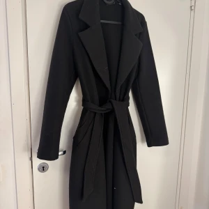 Svart kappa från Vero Moda med bälte - Svart lång kappa från Vero Moda med klassisk krage och knytbälte i midjan. Snygg och stilren design med två fickor framtill och långa ärmar. Perfekt för dig som gillar minimalistisk och clean look.