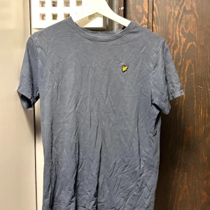 Blå t-shirt från Lyle & Scott Junior - Snygg blå t-shirt från Lyle & Scott Junior med klassisk rund hals och korta ärmar. På bröstet sitter den ikoniska gula fågel-loggan. T-shirten är gjord i mjuk bomull och har en enkel, stilren design som funkar till många olika looks.