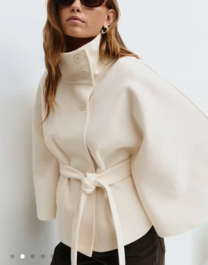 Gina tricot belt cape jacket  - Helt oanvänd, köpt för 3 månader sedan. Säljer en krämvit kappa med hög krage, knappar och knytskärp i midjan. Jackan har vida, trekvartsärmar och en stilren, minimalistisk look. Perfekt för dig som gillar en clean och modern vibe. Materialet känns mjukt och lyxigt.