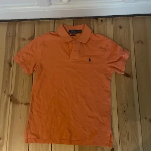 Orange pikétröja från Polo Ralph Lauren - Snygg orange pikétröja från Polo Ralph Lauren i slim fit-modell. Klassisk krage med två knappar och den ikoniska blå logon broderad på bröstet. Perfekt för dig som gillar stilrena och sportiga plagg. Tillverkad i mjuk bomull som är skön mot huden.