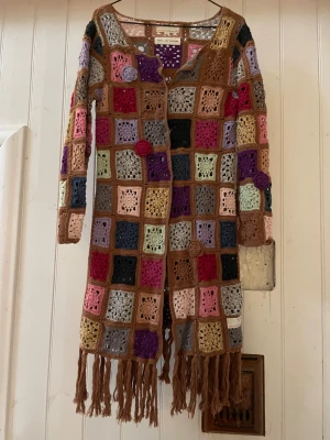 Odd Molly långkofta virkad mormorsrutor - Unik långkofta från Odd Molly, virkad i färgglada rutor med blommiga detaljer och fransar längst ner. Koftan  har lång ärm och är gjord i bomull med inslag av glittertråd. Färgerna är blandade med beige, rosa, lila, röd, svart, grå och guld. Perfekt för dig som gillar boho och virkat. St 1