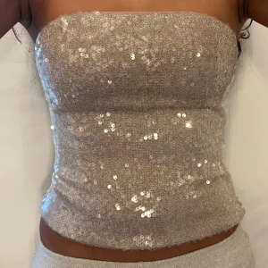 Glittrig tubtopp i champagne från Zara - Säljer en snygg tubtopp från Zara i champagnefärg med glittriga paljetter över hela ytan. Toppen är bandeau-modell, axelbandslös och har en tight passform. Perfekt för att sticka ut med en glittrig vibe. Materialet är syntet och toppen är fodrad på insidan.
