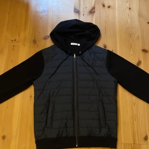 Svart/blå Cardigan från Kappahl - Mörkblå och svart cardigan från Kappahl i storlek M. Jackan har quiltad framsida, mjuka ärmar och rygg i sweatshirt material samt en stor huva med snörning. Dragkedja framtill och ribbade muddar vid nederkant och ärmslut. Perfekt för kyliga dagar. Använd enstaka gånger så den är i utmärkt skick 🤗