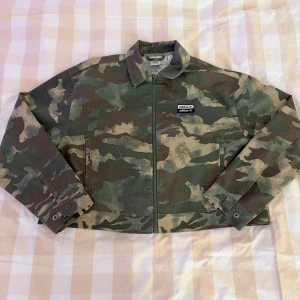 Camouflage overshirt från Adidas - Snygg overshirt från Adidas med klassiskt camouflagemönster i gröna och beige toner. Jackan har dragkedja framtill, krage och två sidofickor. Adidas-logga på bröstet ger en sportig vibe. Perfekt för dig som gillar streetwear och vill sticka ut.