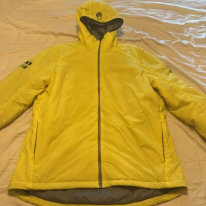 Gul pufferjacka från Sail Racing - Grym gul pufferjacka från Sail Racing med huva och svart logga på ärmen. Jackan har hel dragkedja framtill och två sidofickor. Insidan är grå och materialet känns syntetiskt, perfekt för kalla dagar. Snygg och sportig look med coola detaljer.