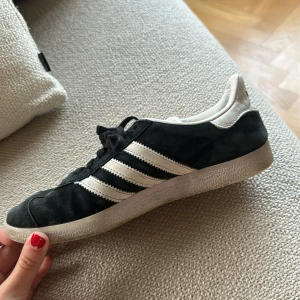 Adidas Gazelle svarta sneakers - Svarta Adidas Gazelle sneakers med klassiska vita ränder på sidorna och vit häl med Adidas-logga. Skorna har snörning och platt sula, tillverkade i mocka och skinn. Snygg retrostil med rund tå och guldtext på sidan.