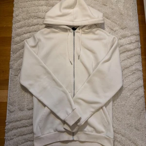 Vit/Beige hoodie med dragkedja från SMOG - Zara - Vit/beige hoodie från SMOG köpt i ZARA i slim fit med huva och snörning. Tröjan har dragkedja framtill, två fickor och ribbade muddar vid ärmslut och nederkant. Tillverkad i mjuk bomull, perfekt för chill dagar. Storlek L passar även storlek M modellen är slim fit.