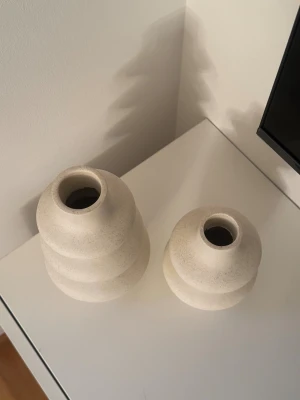 Blomvaser - Bilderna visar två dekorativa vaser i beige keramik med organisk, vågig form. Ytan är matt och lätt prickig, vilket ger ett modernt och stilrent intryck. Båda vaserna ser ut att vara i mycket gott skick utan synligt slitage eller defekter. Perfekt som inredningsdetalj.