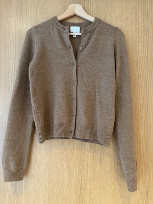 Brun beige stickad kofta från H&M - Finstickad mångsidig cardigan kofta med knappar. Snygg brun beige kofta från H&M i stickad kvalitet, storlek XS-S. Modellen har rund hals, långa ärmar och knappar hela vägen framtill. Perfekt att slänga över en t-shirt eller linne för en chill stilren look. Ribbad kant vid hals, ärmslut och nederkant.