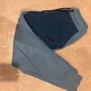 Nike tech blå mjukisbyxor - Nike mjukisbyxor i två nyanser av blått med elastisk midja och svart snörning. Byxorna har ribbade muddar vid bensluten och en svart Nike-logga på låret. Perfekta för chill eller träning, med en loose fit och mjukt material.