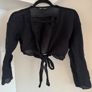 Svart bolerotopp med knyt från Zara - Svart bolerotopp från Zara i tunn, lätt transparent bomull. Modellen är croppad med långa ärmar och har snygg knytning framtill. Perfekt för att lägga till en cool layering-effekt till din outfit. Toppens tyg har en diskret randig struktur som ger extra vibe.