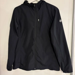 Svart vindjacka från 2117 of Sweden - Svart vindjacka från 2117 of Sweden med huva och diskret logga på ärmen. Jackan har hel dragkedja framtill och en bröstficka med dragkedja. Lätt och smidig design, perfekt för blåsiga dagar. Materialet är syntet och jackan har långärmade ärmar.