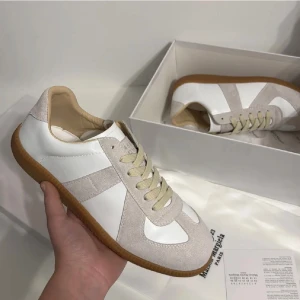 Maison Margiela Replica Gats sneakers vit/beige - Maison Margiela Replica sneakers i vitt och beige med klassisk gummisula. Skorna har snygga mockadetaljer, snörning och rund tå. Materialet är en mix av skinn och mocka, vilket ger en lyxig känsla. Perfekt för dig som gillar stilrena och trendiga sneakers.