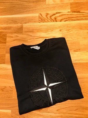 Svart Stone Island sweatshirt S - Svart sweatshirt från Stone Island i storlek S. Tröjan har det klassiska broderade Stone Island-emblemet på ärmen och en stor logga framtill. Mjuk bomullskvalitet och rund hals. Perfekt för dig som gillar streetwear och vill ha en clean look.