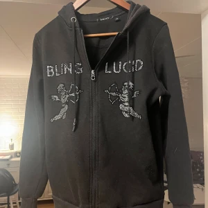 Svart hoodie med strass från Blinglucid - Svart hoodie från Blinglucid med dragkedja, huva och snörning. Framsidan har coola strassdetaljer med texten 'BLING LUCID' och två änglar i strass. Perfekt för dig som gillar streetstyle och bling.