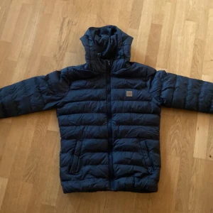 Mörkblå pufferjacka Urban Classics S - Säljer en mörkblå pufferjacka från Urban Classics i storlek S. Jackan har huva, dragkedja framtill och två sidofickor. Den är quiltad och har en snygg Urban Classics-logga på bröstet. Perfekt för kalla dagar när du vill hålla stilen.