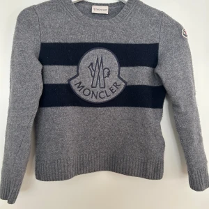 Grå stickad Moncler tröja med logga (Barn) - Snygg grå stickad tröja från Moncler med mörkblå ränder och stor broderad logga på bröstet. Rund hals och långa ärmar, perfekt för dig som gillar exklusiva märken och stilren design. Köpt på NK i Stockholm passar en 7 åring. Nypris 3200:-