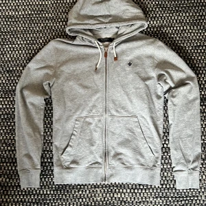 Grå hoodie från Morris med dragkedja - Snygg grå hoodie från Morris med hel dragkedja, huva med vita snören och bruna detaljer. Tröjan har två fickor fram och ett mörkblått broderat emblem på bröstet. Tillverkad i mjuk bomull, perfekt för chill dagar. Passformen är normal.