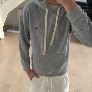 Grå Ralph Lauren Hoodie - Snygg grå hoodie från Polo Ralph Lauren med klassisk logga på bröstet. Tröjan har huva med vita snören, ribbade muddar och en stor magficka. Perfekt för chill dagar och enkel att matcha med jeans eller mjukisbyxor. HELT NY MED PRISLAPP. Passar även XS.