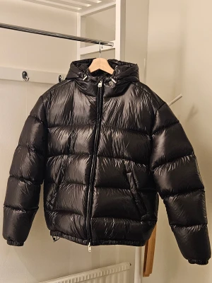 Svart pufferjacka från Zara - Säljer en svart pufferjacka från Zara i storlek M. Jackan har en glansig finish, huva med snörning och dragkedja framtill. Fylld med syntetmaterial och har två sidofickor. Perfekt för kalla dagar och har en riktigt chill oversized vibe. Nypris 1400 kr