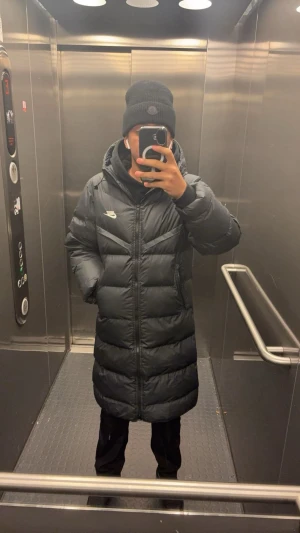 Svart lång Nike pufferjacka XS - Lång svart pufferjacka från Nike med Storm-FIT-teknologi. Jackan har huva, dragkedja framtill och diskret Nike-logga på bröstet. Fylld med syntetmaterial för extra värme och har en clean, sportig look. Perfekt för kalla dagar.