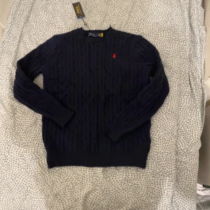 Mörkblå stickad tröja Polo Ralph Lauren - Snygg mörkblå stickad tröja från Polo Ralph Lauren med klassiskt kabelstickat mönster och röd broderad logga på bröstet. Rund hals och långa ärmar, perfekt för dig som gillar stilren och tidlös design.