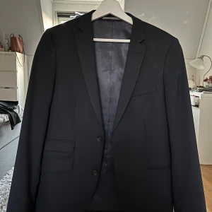 Svart kavaj från Riley Slim Fit - Stilren svart kavaj från Riley i slim fit-modell. Klassisk design med två knappar framtill, flera fickor och snyggt foder. Perfekt för dig som gillar en modern och elegant look. Materialet är troligtvis ullblandning med polyesterfoder.