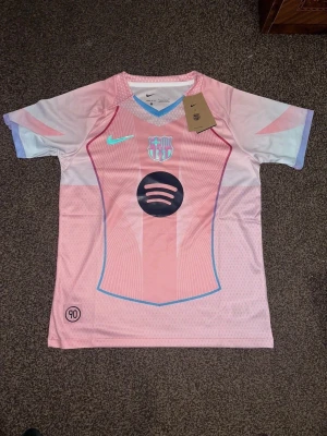 Rosa FC Barcelona fotbollströja Nike - Säljer en FC Barcelona fotbollströja från Nike i en snygg ljusrosa färg med vita och blå detaljer. Tröjan har korta ärmar, klubbmärke på bröstet och Spotify-logga i svart. Materialet är lätt och andas, perfekt för träning eller match.