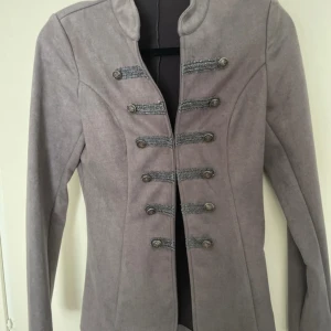 Grå rock med dekorativa knappar - Snygg grå rock med militärinspirerade detaljer och dekorativa silverfärgade knappar framtill. Jackan har lång ärm, figurnära passform och stårkrage. Materialet känns mjukt och har en mockaliknande yta. Perfekt för dig som gillar stilrena och unika plagg.