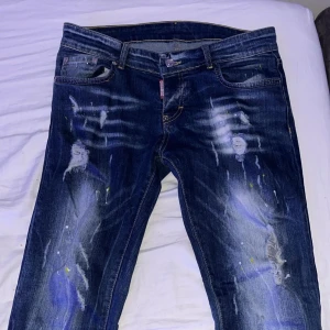 Dsquared2 blå jeans med slitningar - Snygga blå jeans från Dsquared2 med coola slitningar, färgstänk och distressed detaljer både fram och bak. Klassisk femficksmodell med gul kontrastsöm och Dsquared2-logga på bakfickan och vid gylfen. Perfekt för dig som gillar streetstyle och unik look.