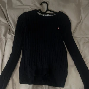 Svart stickad tröja Polo Ralph Lauren - Svart kabelstickad tröja från Polo Ralph Lauren med lång ärm och rund hals. Klassisk rosa broderad logga på bröstet. Tröjan har ribbade muddar och är gjord i mjuk bomull, perfekt för dig som gillar stilrena och tidlösa plagg.