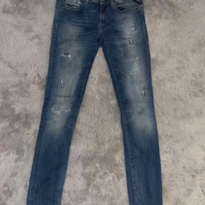 Replay blå jeans med slitningar - Tvärfeta  blå jeans från Replay i anbass modellen med coola slitningar och tvättade detaljer. Klassisk femficksmodell med knapp och dragkedja.