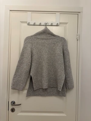 Grå ribbad polotröja i ull - Säljer en grå polotröja med ribbad struktur och lång ärm. Tröjan har en hög krage och slitsar på sidorna vilket ger en chill och avslappnad vibe. Perfekt för kalla dagar och enkel att matcha med jeans eller leggings.