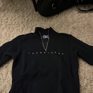 Svart half zip tröja Jack & Jones - Svart långärmad tröja från Jack & Jones med half zip och ribbad krage. Snyggt tryck med loggan på bröstet. Perfekt för dig som gillar stilrena och enkla plagg. Materialet känns mjukt och skönt mot huden. Mycket bra skick 