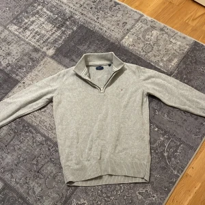 Ljusgrå stickad half zip-tröja GANT - Snygg ljusgrå stickad tröja från GANT med half zip och ribbad krage. Klassisk design med broderad logga på bröstet och långa ärmar. Perfekt för dig som gillar stilrena och bekväma plagg. Passar dig som är 13-14 år.