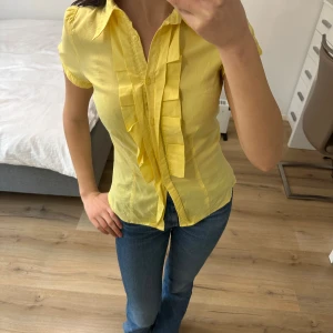 Gul blus med volang och knappar - Snygg gul blus med korta puffärmar och volangdetaljer längs knappraden framtill. Skjortan har klassisk krage och är figurnära i modellen. Perfekt för dig som gillar färg och vill sticka ut lite extra.