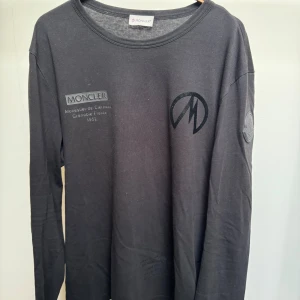 Svart långärmad t-shirt från Moncler - Svart långärmad t-shirt från Moncler med tryck på bröstet och logga på ärmen. Snygg rund halsringning och stilren design. Tillverkad i mjuk bomull som känns skön mot huden. Perfekt för dig som gillar exklusiva streetwear-plagg.