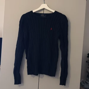 Mörkblå stickad tröja från Polo Ralph Lauren - Snygg mörkblå stickad tröja från Polo Ralph Lauren med långärmad passform och ribbad struktur. Tröjan har en röd broderad logga på bröstet och är perfekt för dig som gillar klassisk stil med en twist.