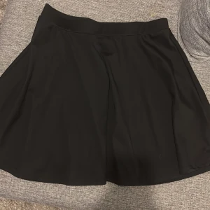 Svart kortkjol med utsvängd form - Säljer en svart kortkjol med snygg utsvängd siluett. Kjolen har en enkel och stilren design utan mönster eller detaljer, vilket gör den lätt att matcha med olika toppar. Materialet känns mjukt och har ett lätt fall. Perfekt för dig som gillar minimalistisk stil.