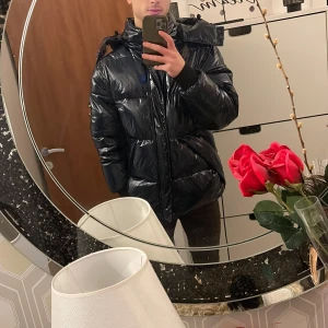 Blå pufferjacka från SikSilk - Säljer en blå, glansig pufferjacka från SikSilk med stor huva och dragkedja framtill. Jackan har quiltad design, lång ärm och coolt emblem på armen. Perfekt för dig som vill ha en varm och snygg jacka med streetkänsla. Fråga om du vill veta mer!. DEN ÄR TYP HELT NY. PRISET KAN DISKUTERAS 