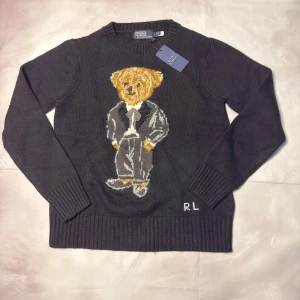 Svart stickad tröja Polo Bear Ralph Lauren - Svart stickad tröja från Polo Ralph Lauren med ikoniska Polo Bear-motiv på bröstet. Rund hals, ribbade muddar och RL-broderi vid nederkanten. Tröjan är långärmad och har en klassisk, normal passform. Perfekt för dig som gillar statement-plagg och snygga detaljer.