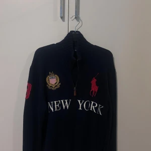 Mörkblå stickad kofta Polo Ralph Lauren - Snygg mörkblå stickad kofta från Polo Ralph Lauren med broderad New York-text och emblem på bröstet, samt röd logga och siffra på ärmen. Koftan har ribbad krage och dragkedja framtill. Perfekt för dig som gillar klassisk college-stil.