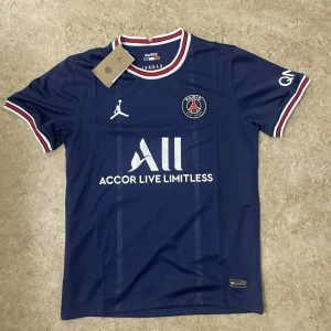 PSG marinblå fotbollströja Jordan - Snygg marinblå PSG fotbollströja från Jordan med vit logga och sponsortryck. Röd, vit och blå ribbad krage och ärmslut. Paris Saint-Germain-emblem på bröstet. Tillverkad i lätt och ventilerande material, perfekt för match eller träning.