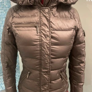 Brun pufferjacka Rockandblue, st 34 - Snygg brun pufferjacka från Rockandblue med huva och flera dragkedjeförsedda fickor. Jackan har quiltade detaljer på axlarna och ett Rockandblue-märke på ärmen. Passformen är figurnära och materialet är syntet, perfekt för kalla dagar. St 34