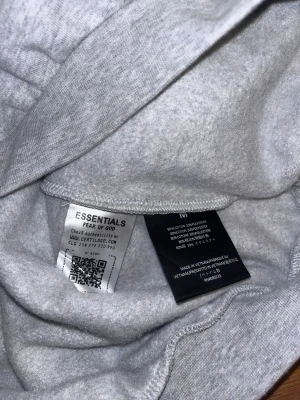 Grå Essentials Fear of God hoodie - Snygg grå hoodie från Essentials Fear of God med svart logotyptryck på bröstet och ryggen. Tröjan har huva, långa ärmar och ribbade muddar. Tillverkad i mjuk bomull och polyesterblandning som känns skön mot huden. Perfekt för dig som gillar streetwear och clean stil.